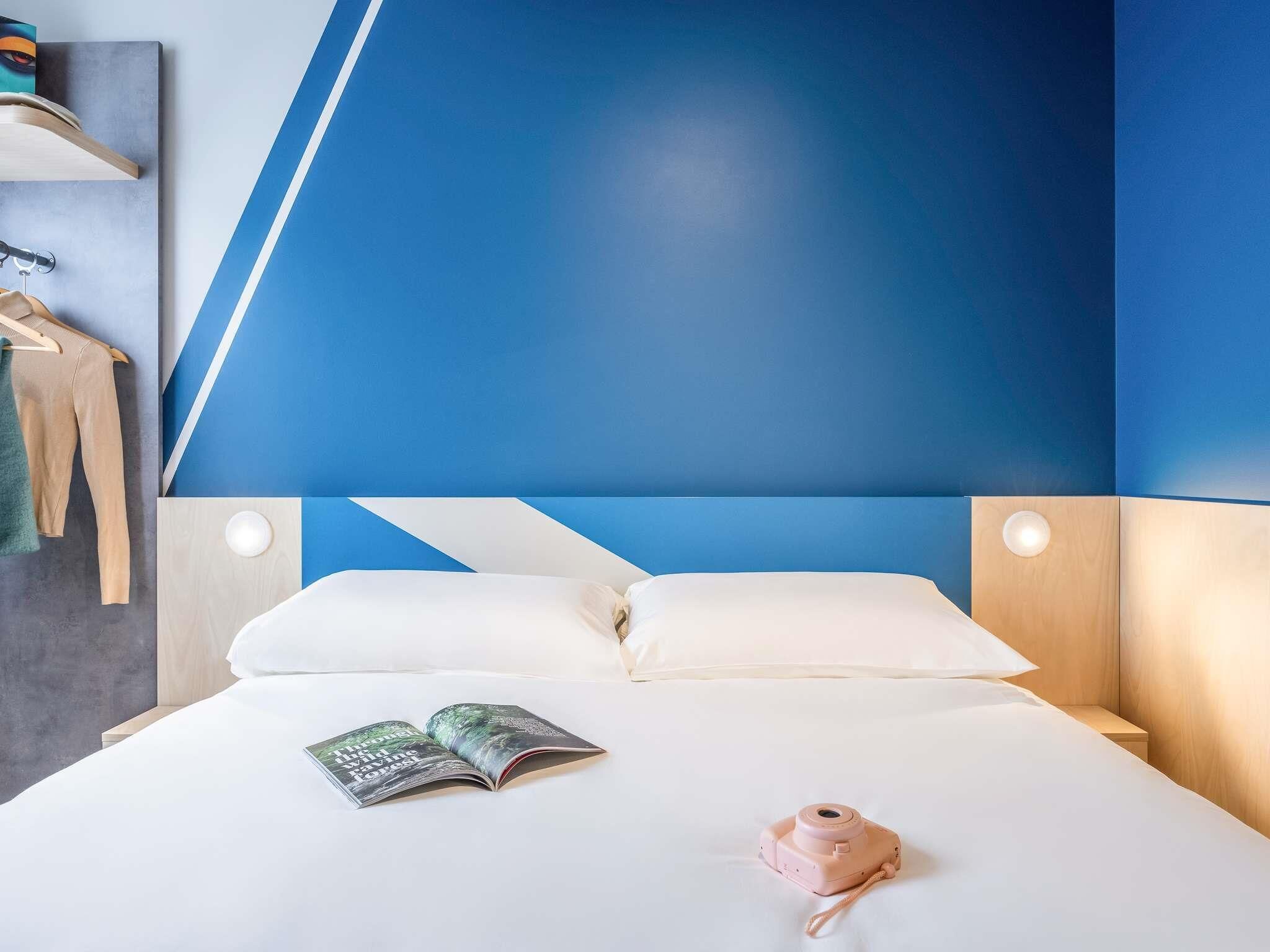 Un lit bien fait, avec du linge de lit blanc, un appareil photo rose et un livre posé sur le lit.