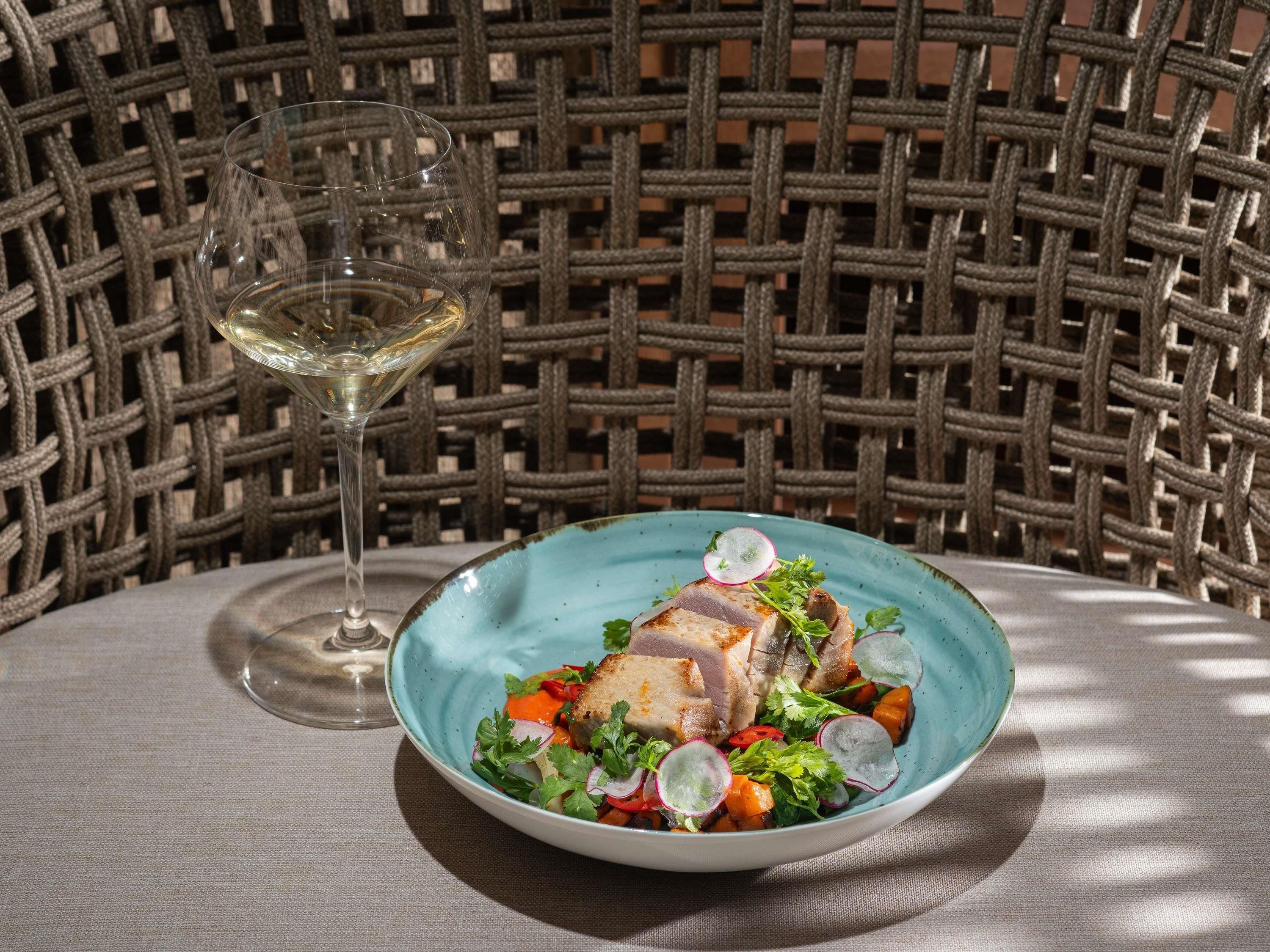 A plate of food with a glass of white wine on a table with a woven chair in the background.