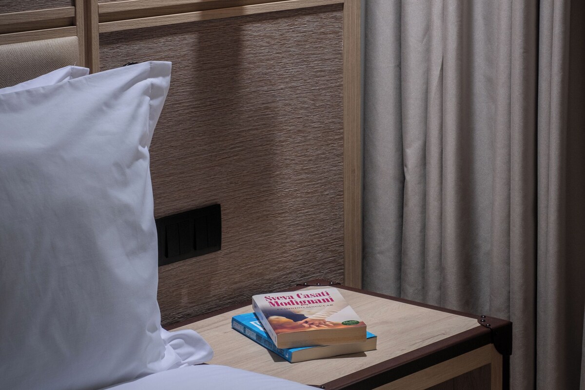Una habitación de hotel con una cama, una mesita de noche con un libro, una lámpara de pared y una cortina.