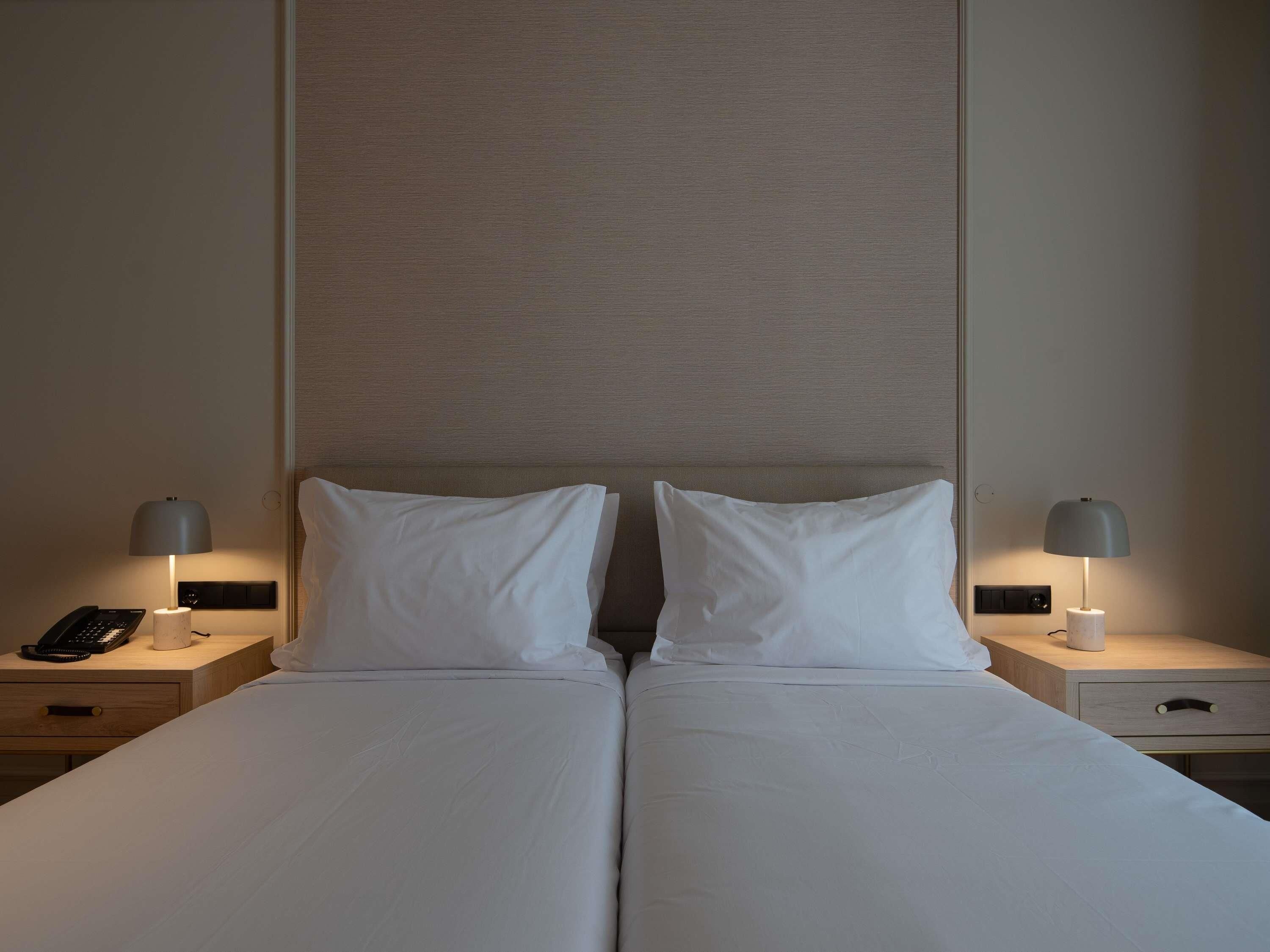 A double bed with white linens, two bedside tables with lamps, a phone, and a wall panel.