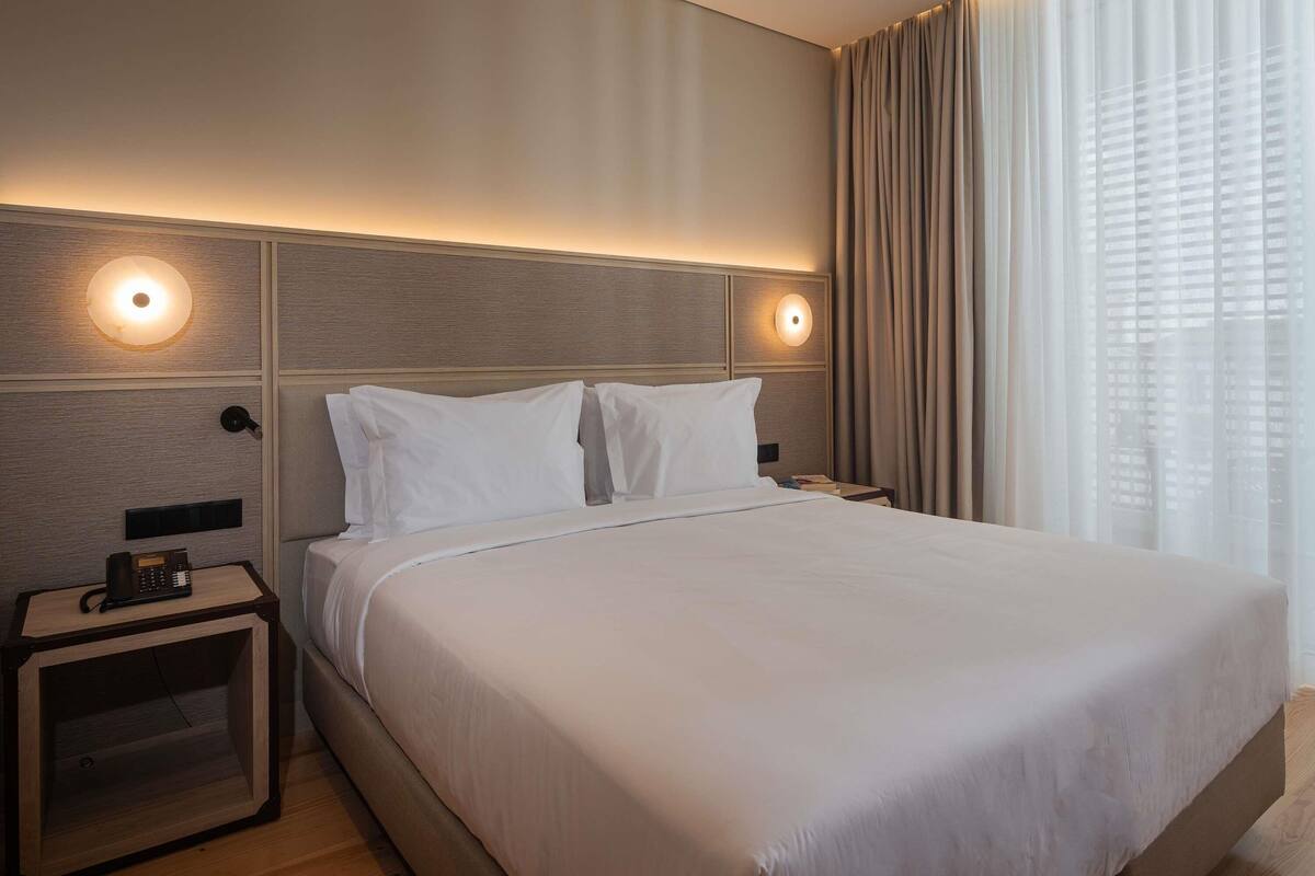 A hotel room with a large bed, two bedside tables, a phone on one table, and a window with curtains.