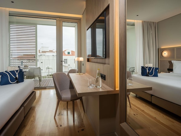 A hotel room with a wooden floor, a dining table, two beds, a TV, and a balcony with a view of buildings.