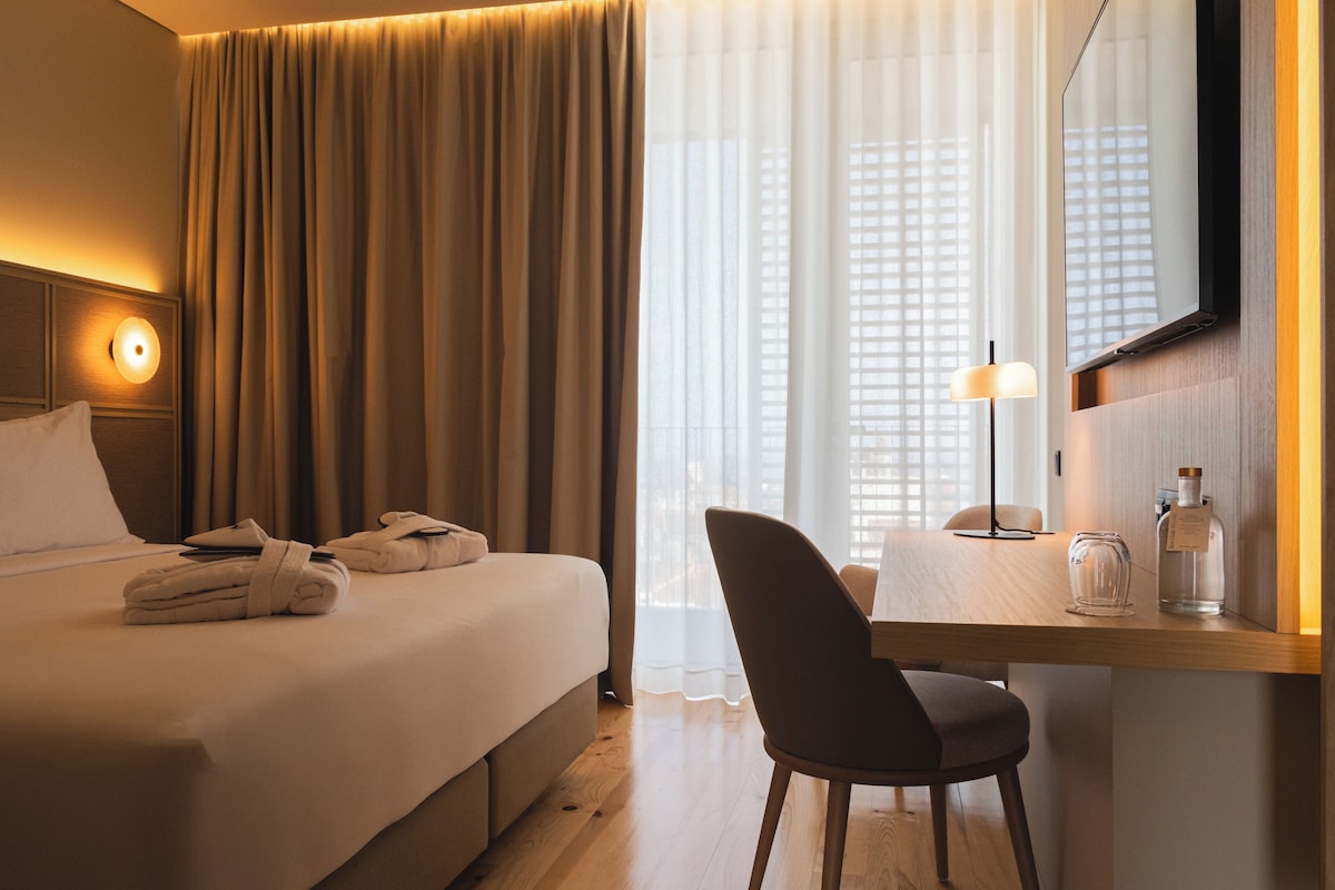 Habitación de hotel con cama, escritorio con silla, espejo y ventana con cortinas.