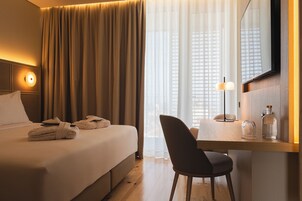 A hotel room with a bed, a desk with a chair, a mirror, and a window with curtains.