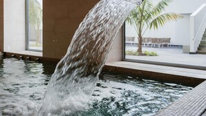 Un elemento d'acqua con cascata in zona piscina, con vista su una palma e area relax all'aperto sullo sfondo.