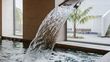 Ett vattenspel med en vattenfallsfunktion i ett poolområde, med utsikt över en palm och utomhusmöbler i bakgrunden.