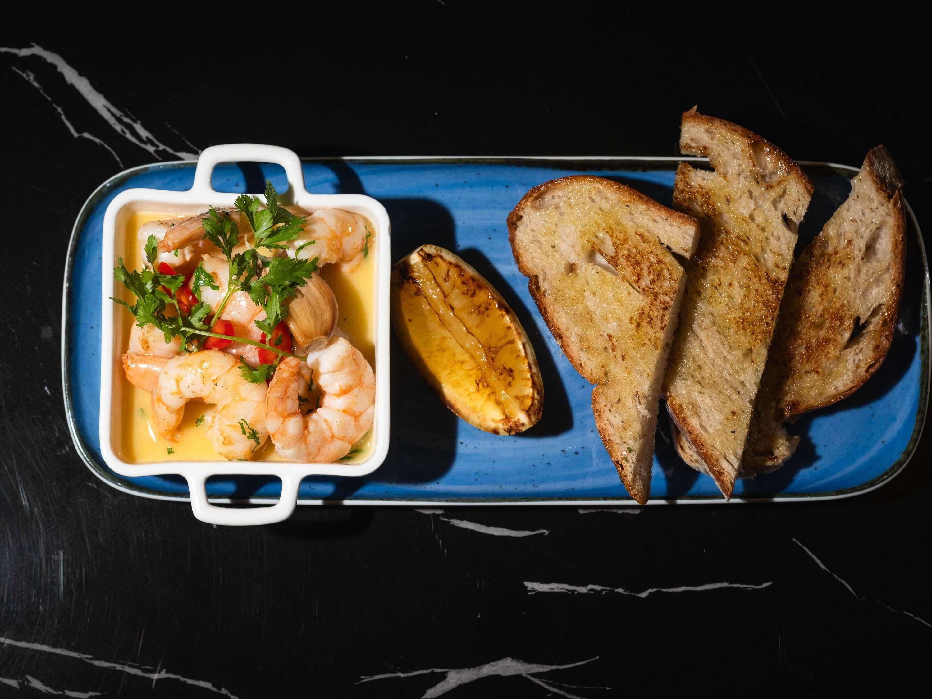 A rectangular dish with shrimp and a lemon half, accompanied by toasted bread slices on a blue plate.