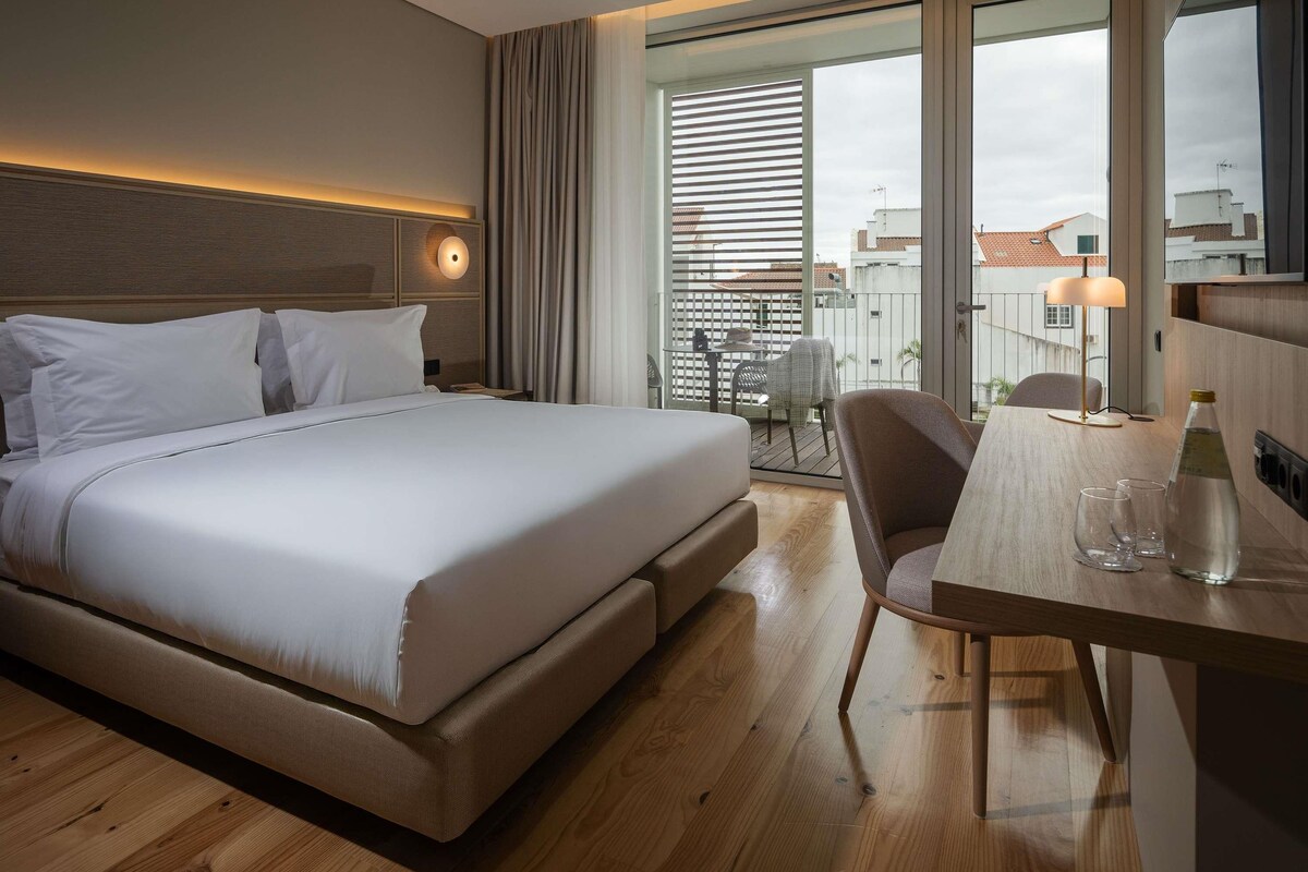 A hotel room with a large bed, a desk with a chair, a table with a lamp, and a view of the city through large windows.
