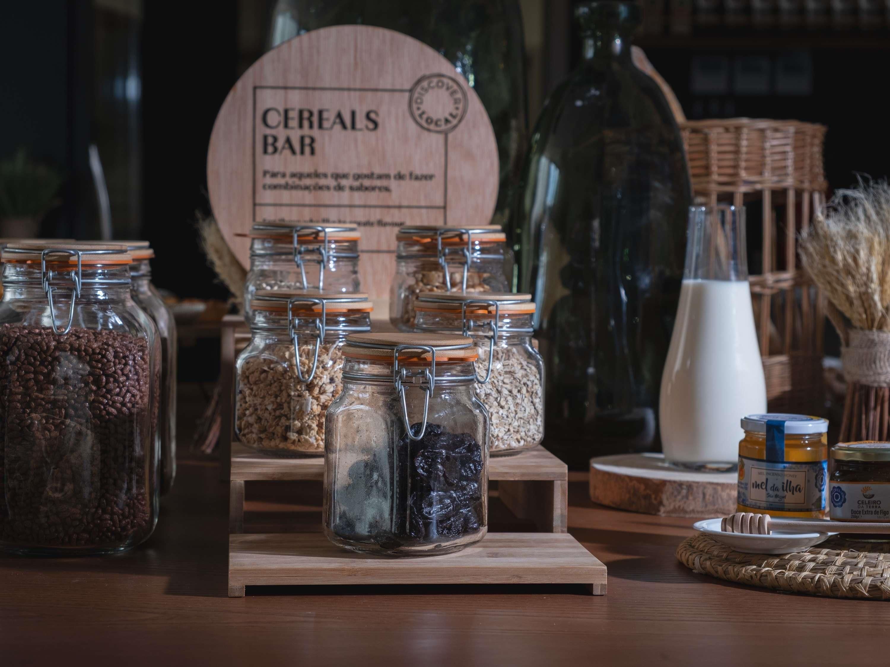 A display of various grains and seeds in glass jars with metal lids, arranged on a wooden stand.