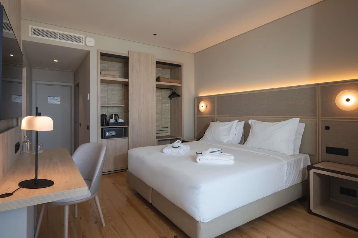 A modern hotel room with a bed, bedside tables, a desk, a chair, and a wardrobe.