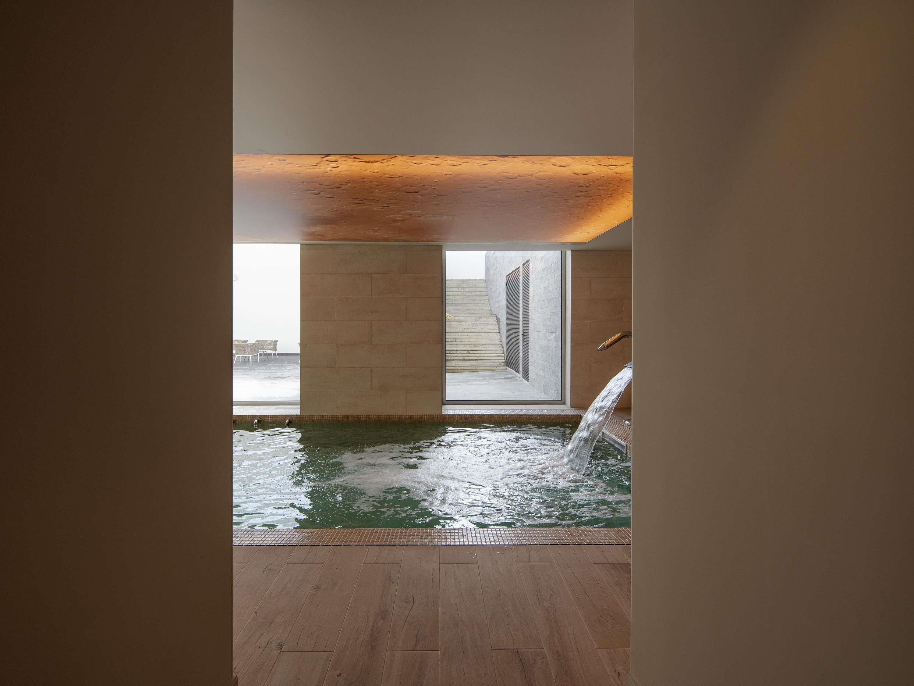 Piscina interior moderna com vista clara para o exterior, piso de madeira e um elemento aquático.