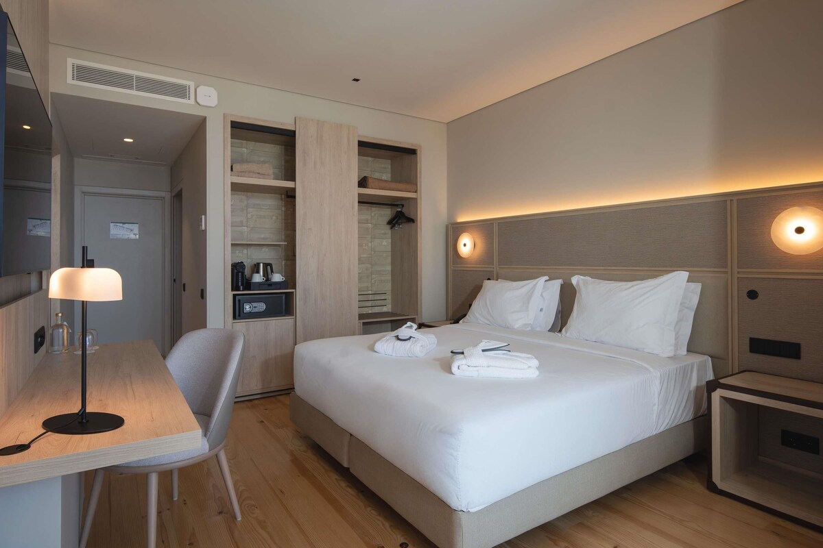 A modern hotel room with a bed, bedside tables, a desk, a chair, and a wardrobe.