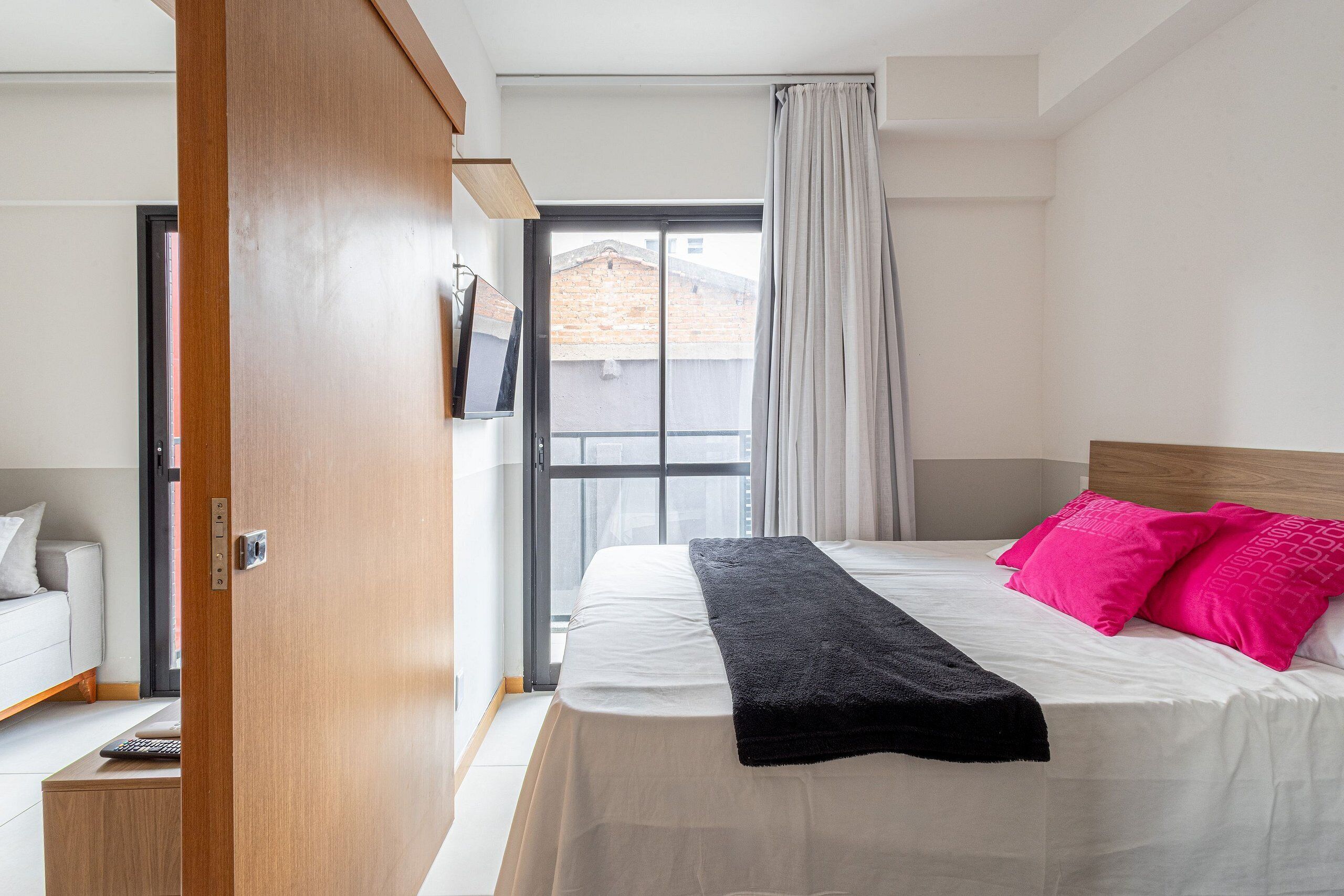 A bedroom with a bed, a wooden door, a window with curtains, and a view of buildings outside.