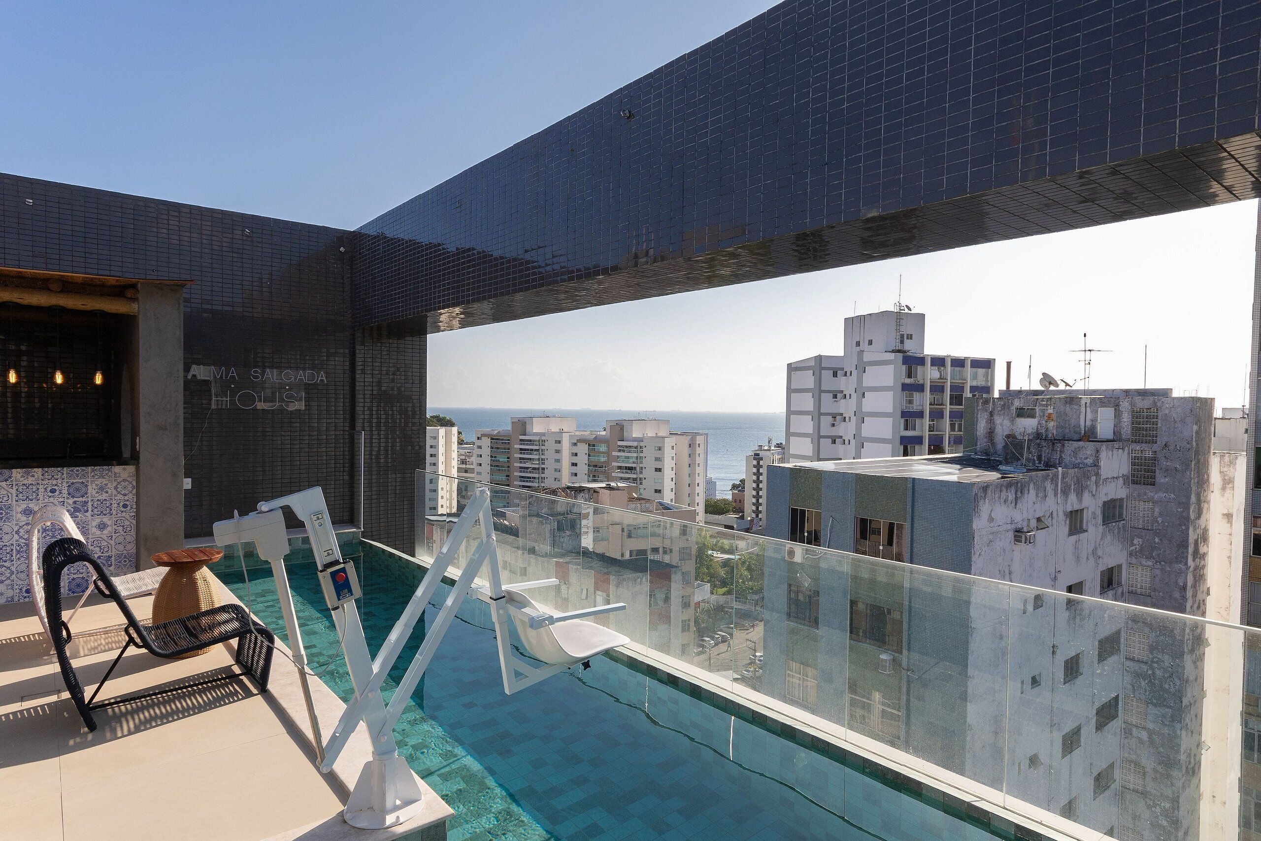 Una piscina en la azotea con balaustrada de cristal, una silla de descanso y vistas a la ciudad y al océano.