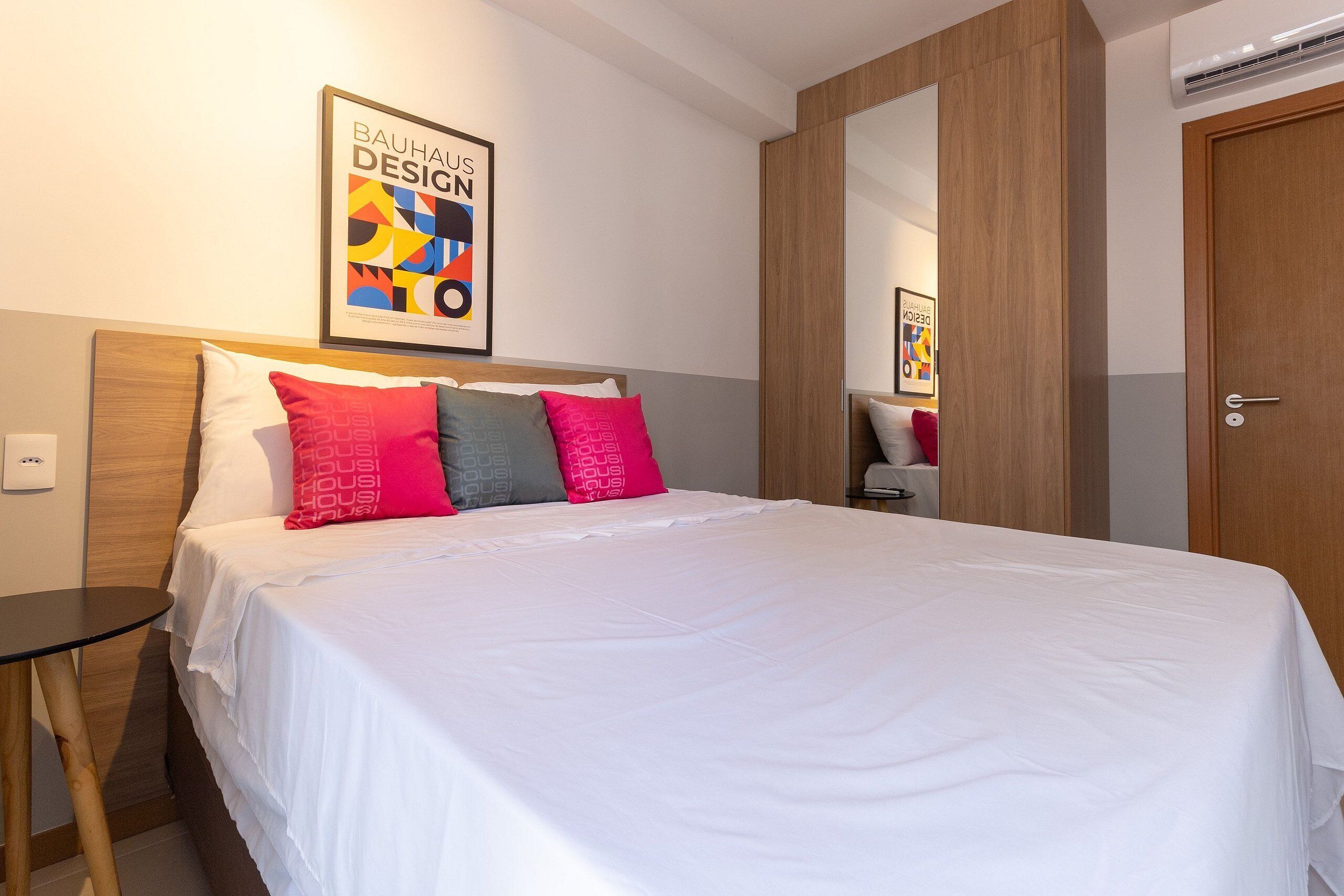 A neatly made bed with white bedding and multiple pillows, a wooden headboard, a small table, and a framed poster on the wall.