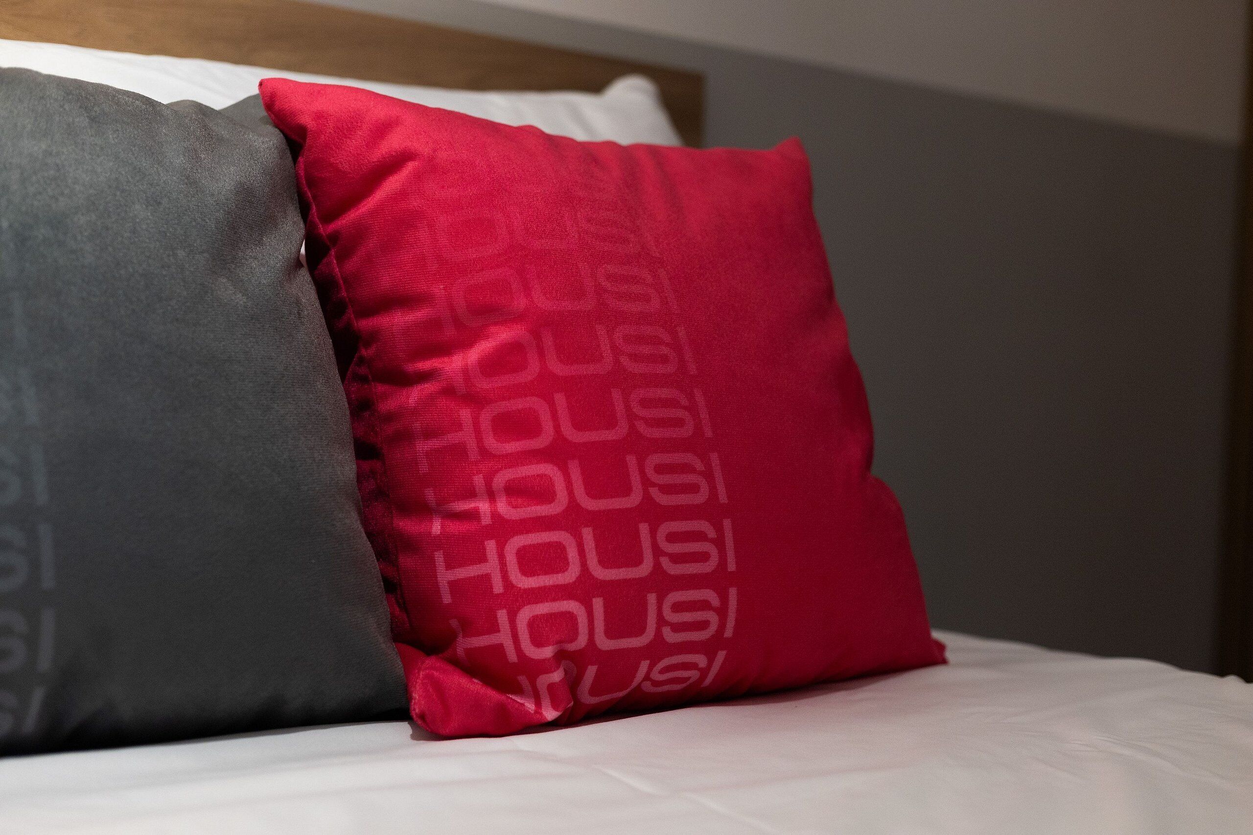 A bed with two pillows, one red and one grey, with the word 'HOUSI' printed on them.