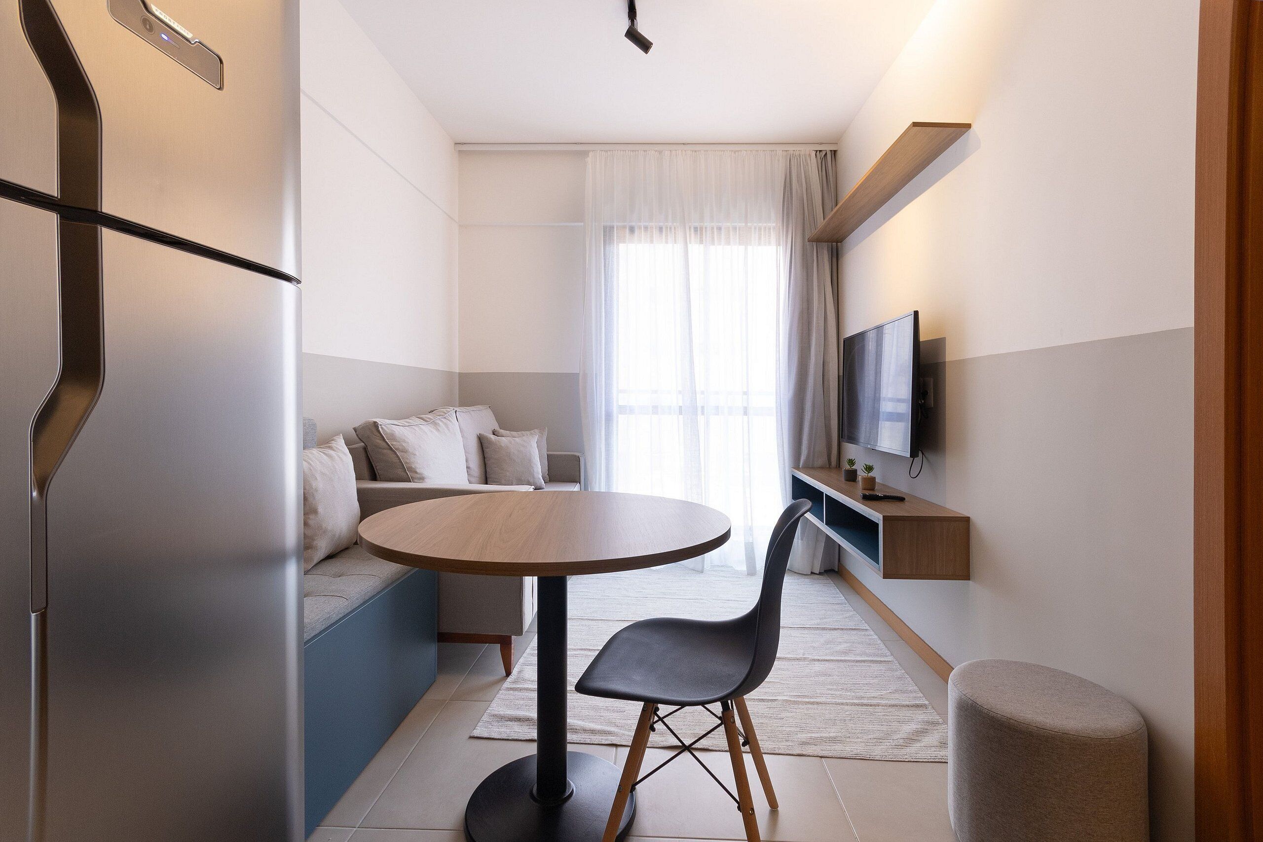A modern hotel room with a round table, a sofa, a TV, and a small wooden shelf.