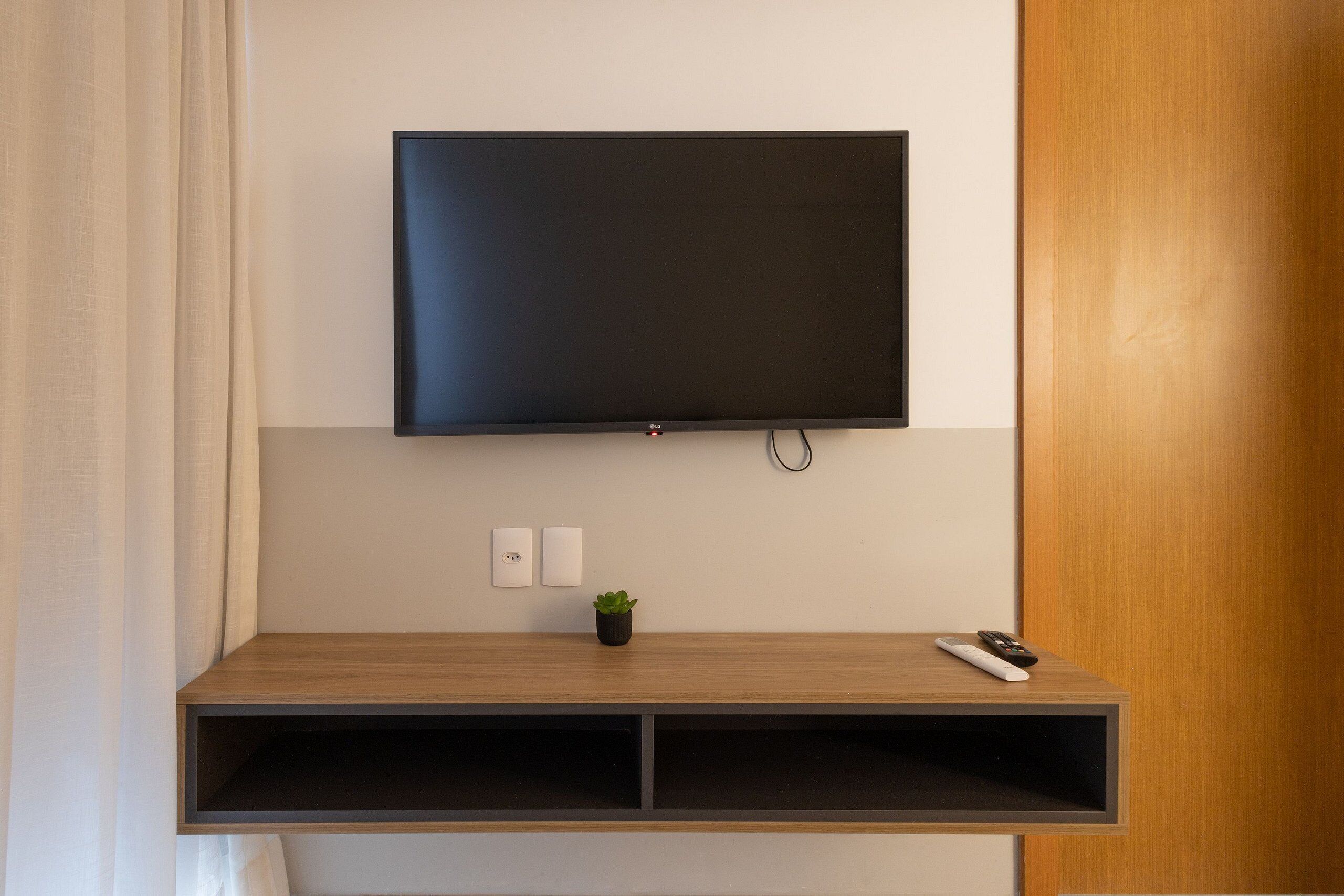 A flat-screen TV mounted on a wall above a wooden console with a small plant and a remote control.