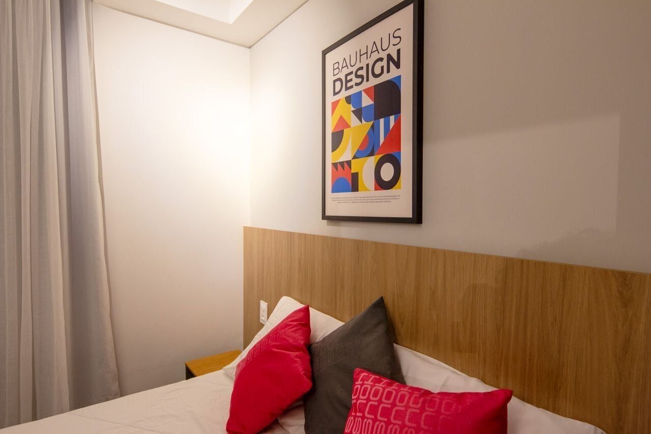 A hotel room with a wooden headboard, a bed with white bedding and red and gray pillows, and a framed Bauhaus Design poster on the wall.