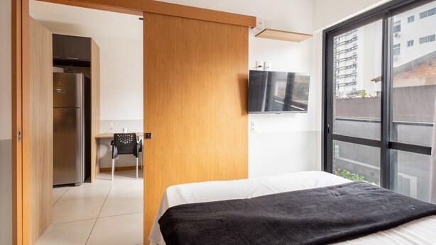 Una habitación de hotel moderna con una cama, un televisor montado en la pared, una puerta corredera de madera y vistas a la ciudad a través de amplios ventanales.