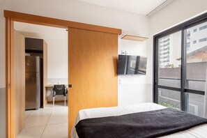 Una habitación de hotel moderna con una cama, un televisor montado en la pared, una puerta corredera de madera y vistas a la ciudad a través de amplios ventanales.