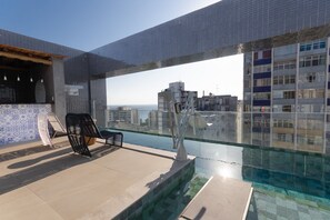 A rooftop pool with a view of the cityscape, a lounge area with chairs, and a tiled wall.