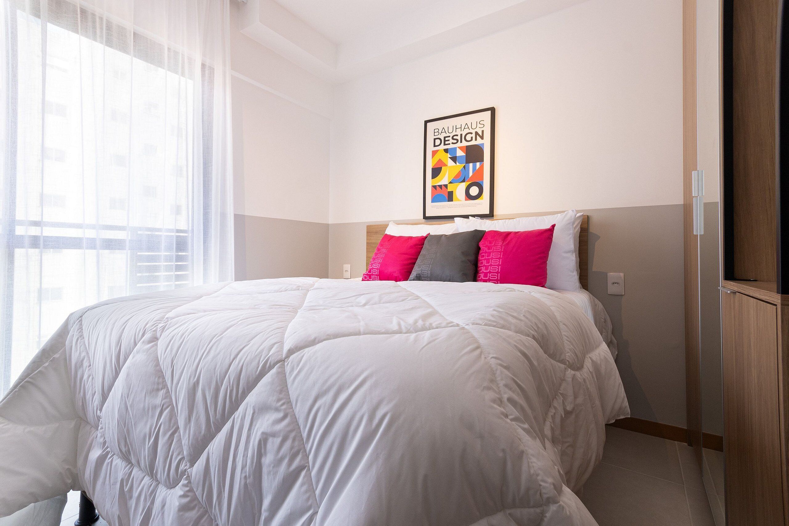 A bedroom with a large bed, a wall-mounted poster, and a window with sheer curtains.