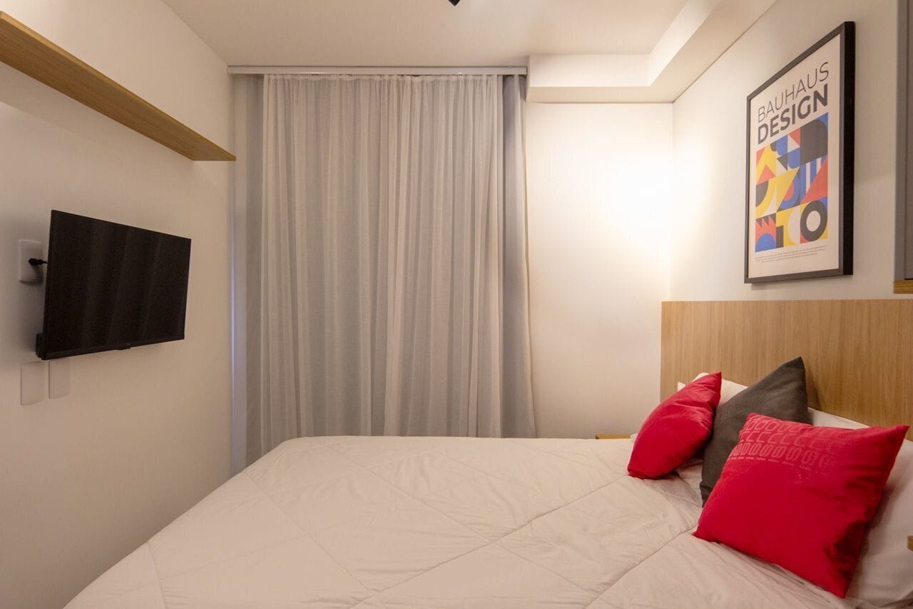 A hotel room with a bed, a flat-screen TV, a framed poster, and a window with curtains.