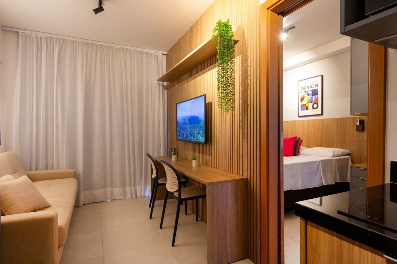 A modern living room with a wooden accent wall, a flat-screen TV, a dining area with a wooden table and chairs, and a bed with a red pillow.