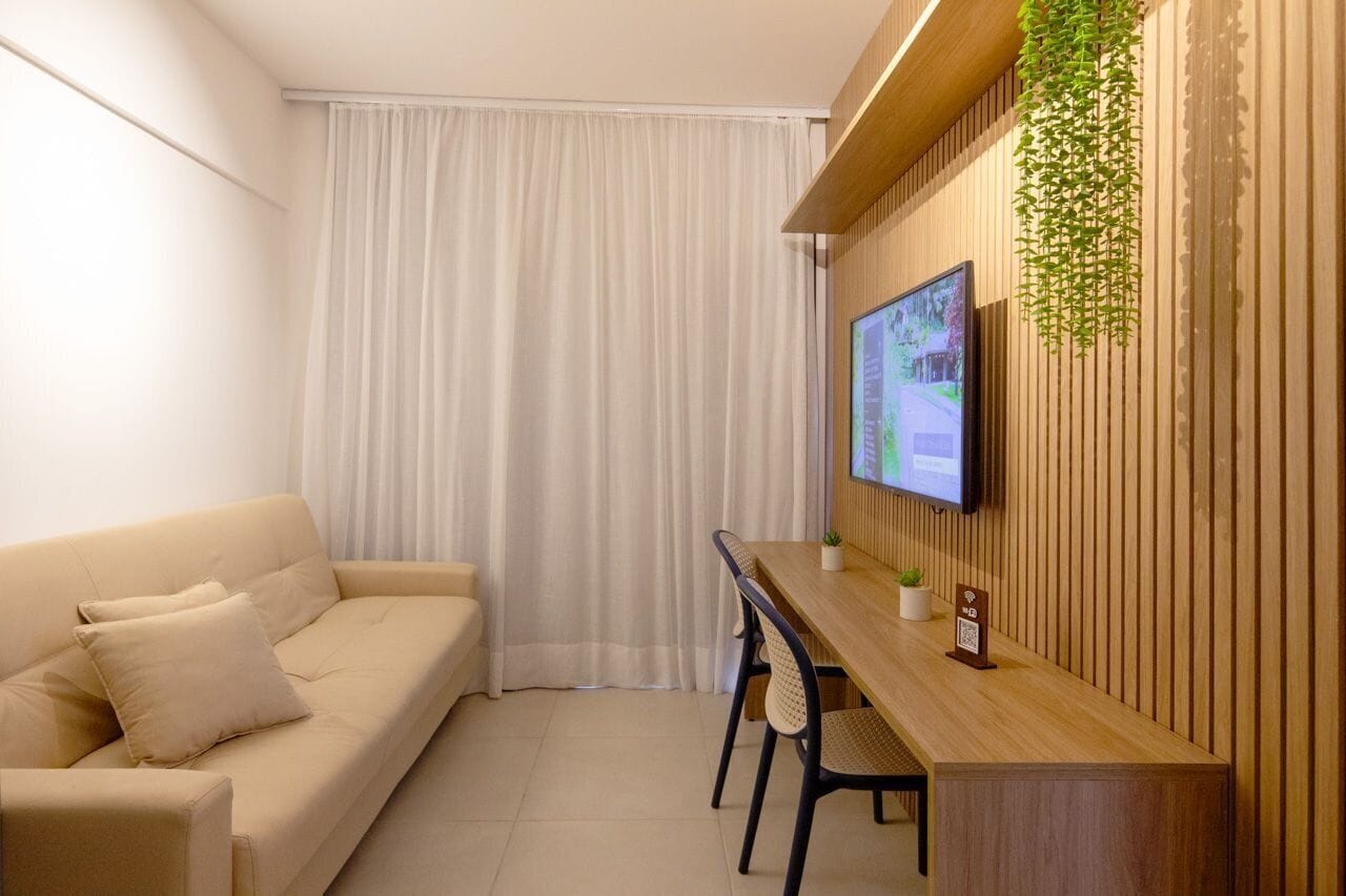 A modern living room with a sofa, a wooden dining table, chairs, a TV mounted on the wall, and a hanging plant.