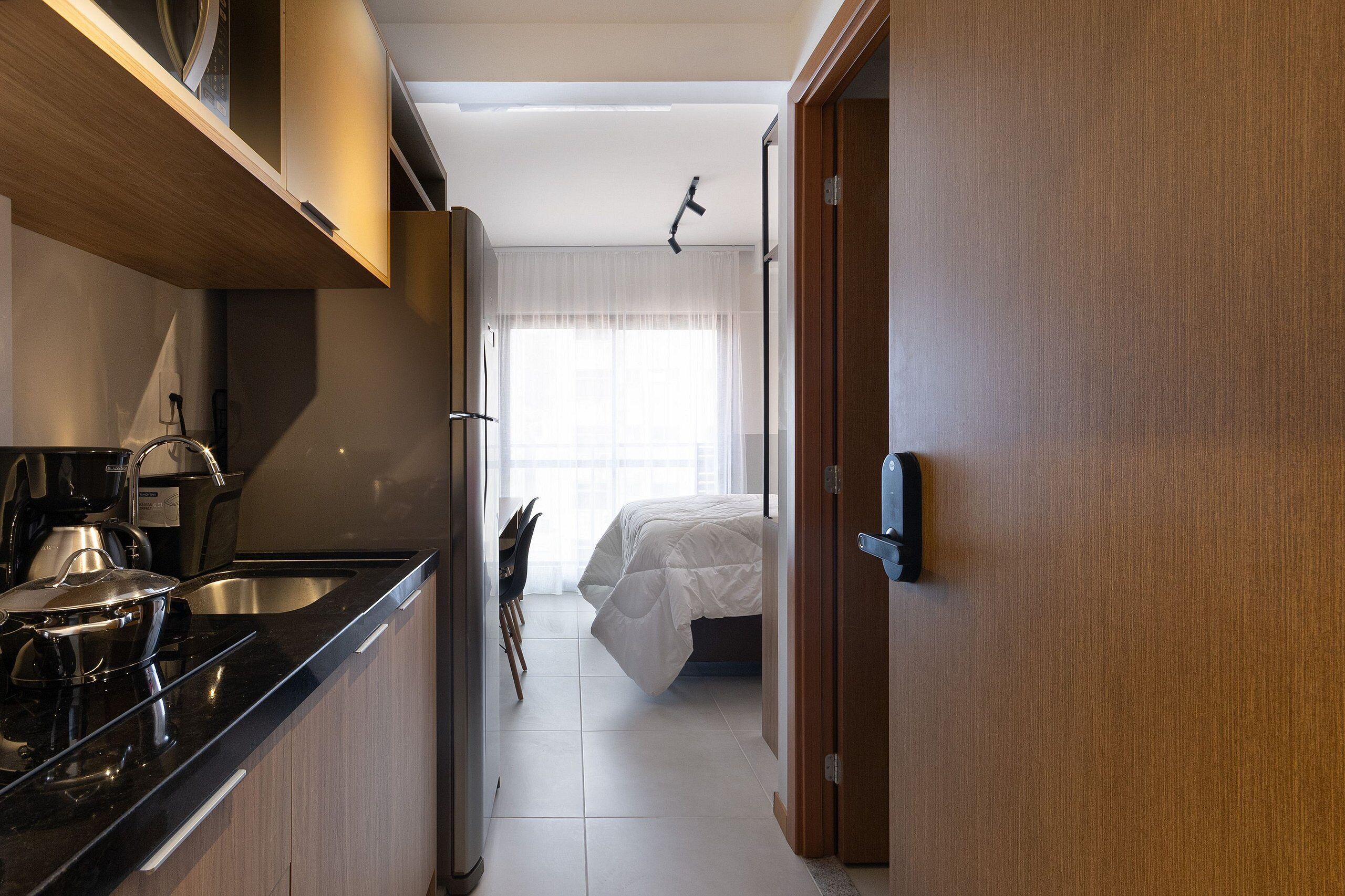 A compact kitchen with modern appliances, a dining area with a chair, and a bed with a white comforter.