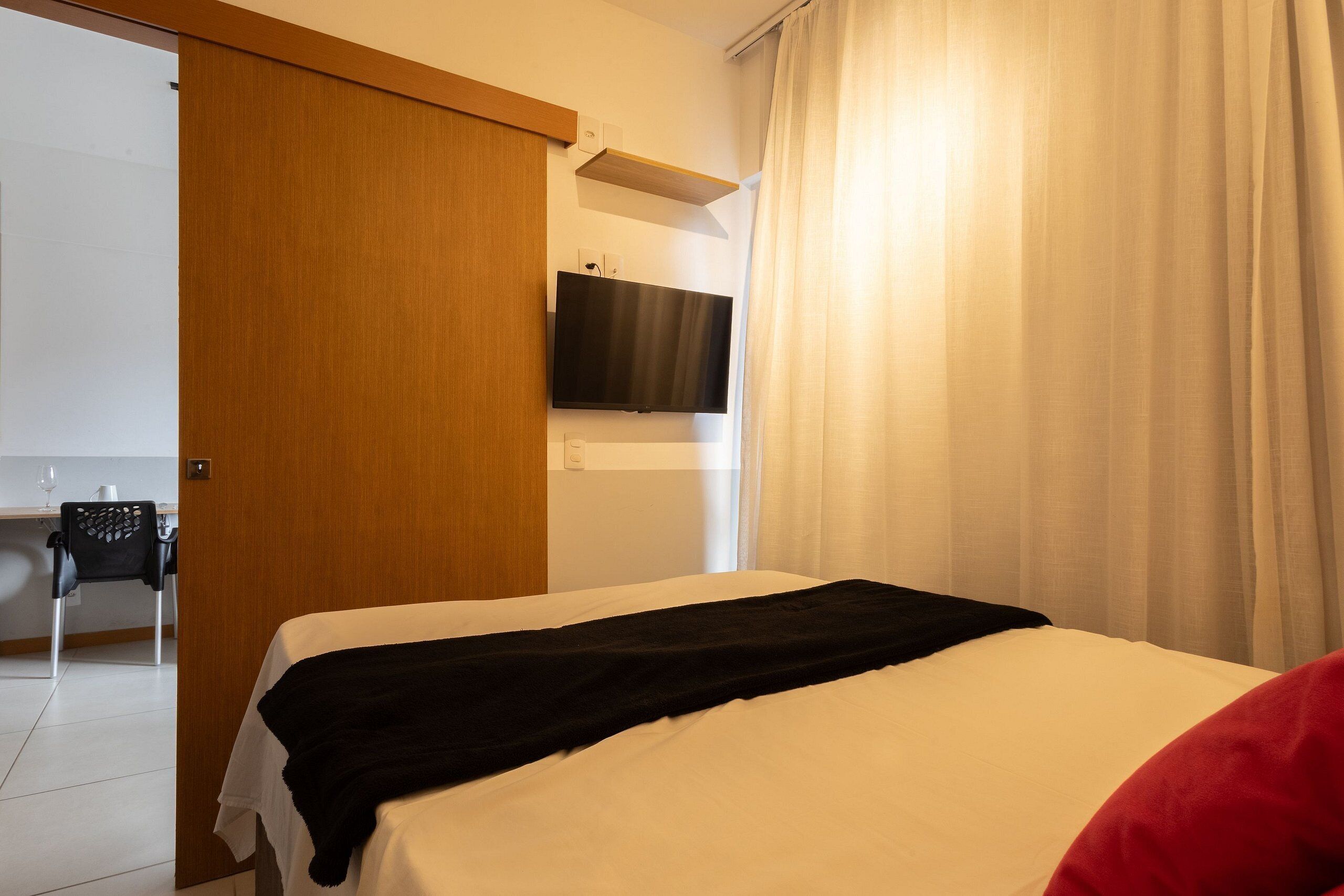 A hotel room with a bed, a TV mounted on the wall, a wooden door, and a window with curtains.