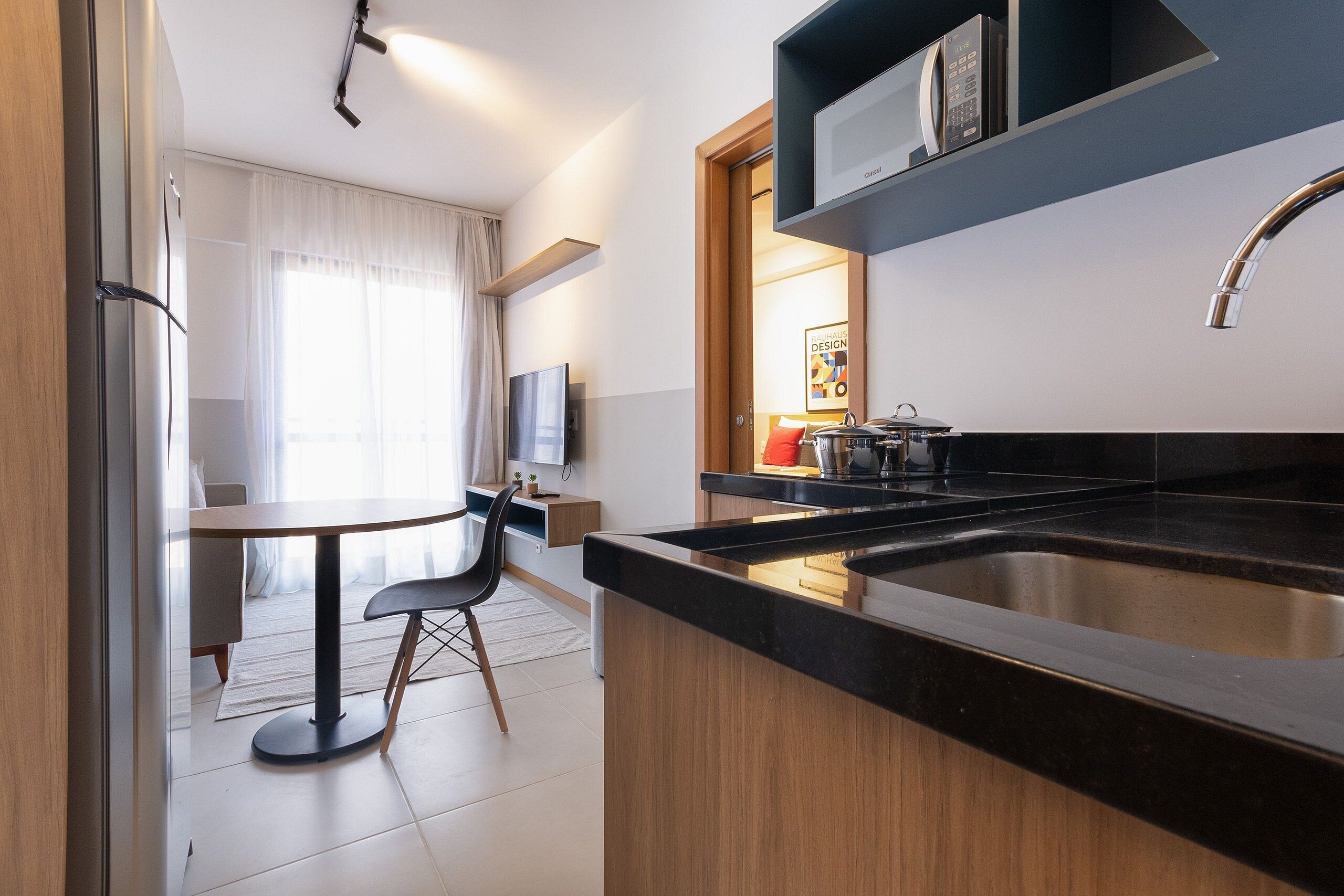 A modern kitchen with a black countertop, stainless steel sink, and a built-in oven. There is a dining area with a table and chairs, and a TV mounted on the wall.