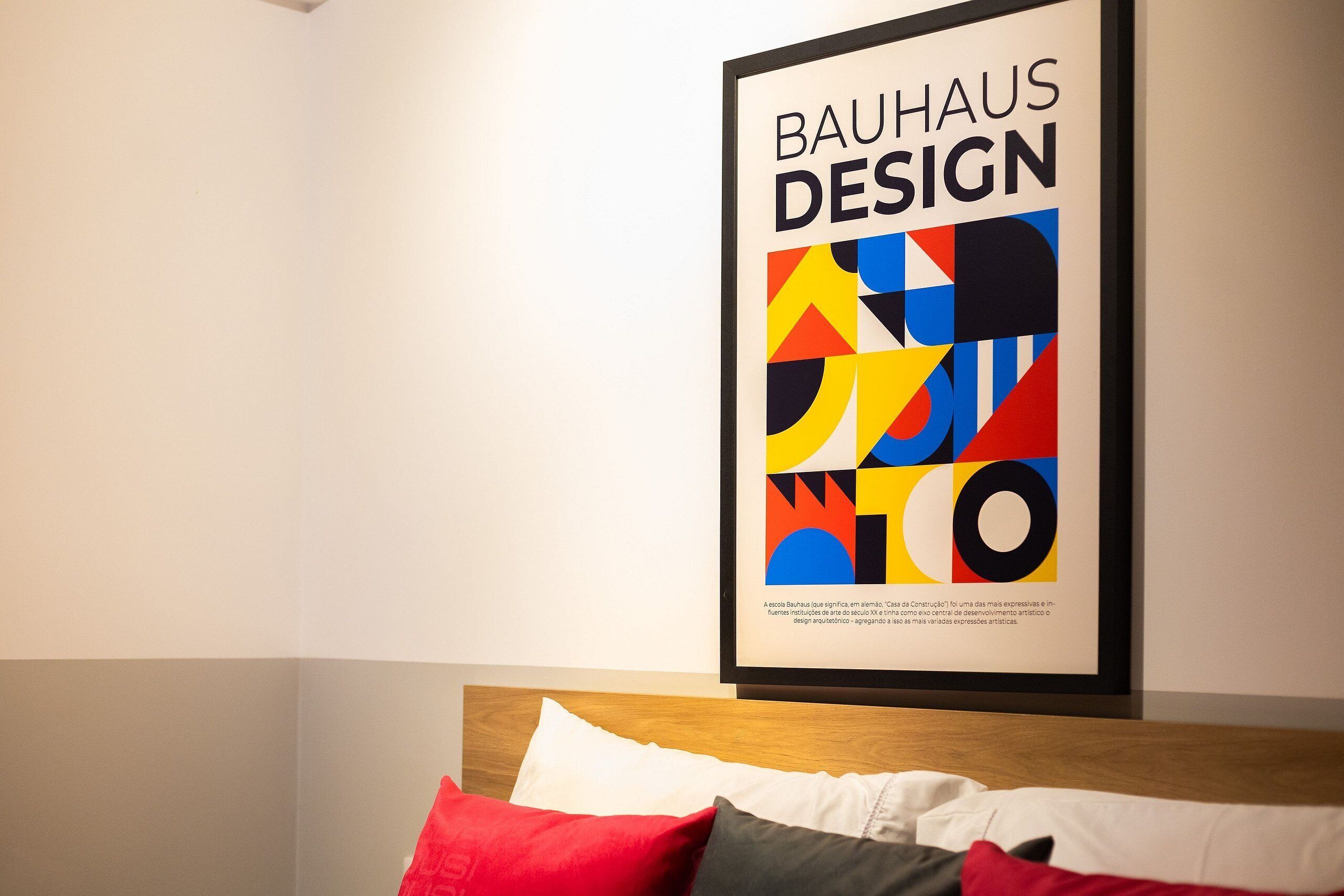 En inramad affisch med en Bauhaus-design, hängd på en vägg ovanför en säng med kuddar.