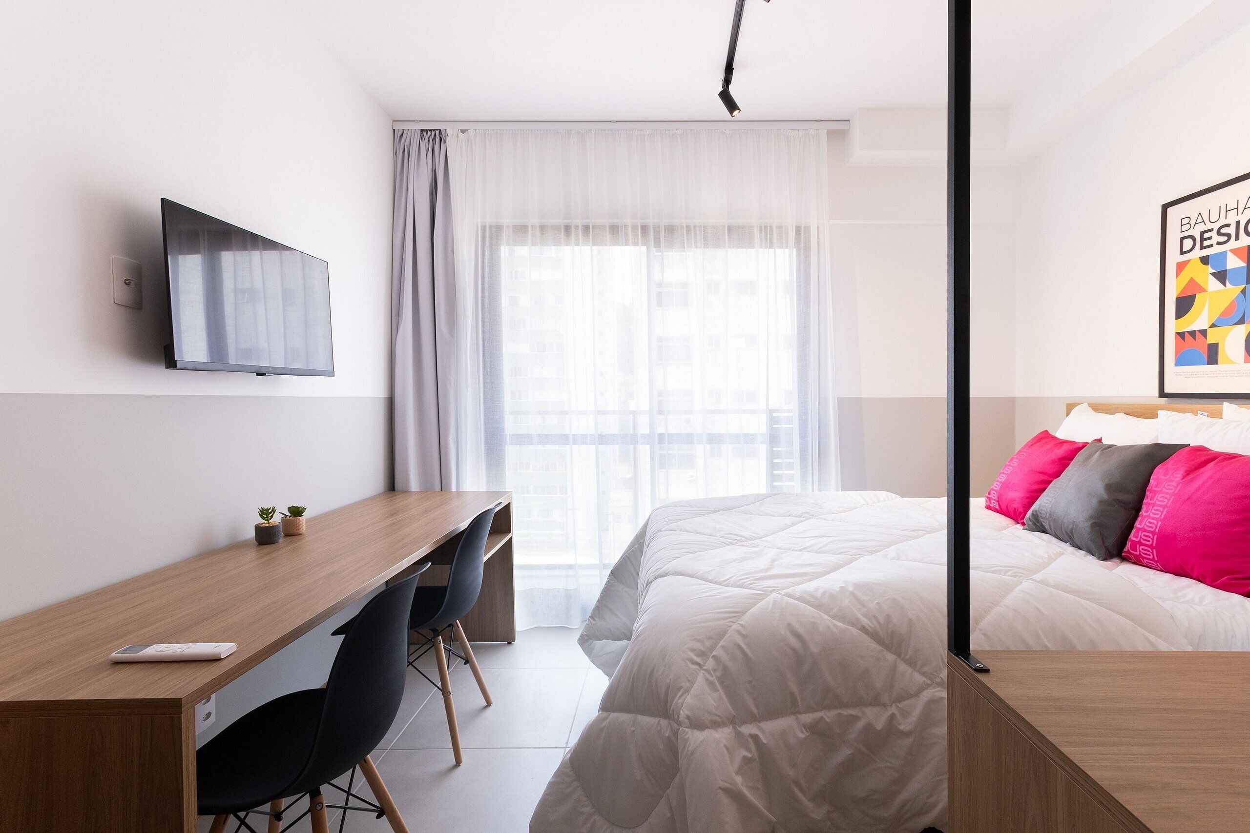 Una habitación de hotel moderna con una cama, un escritorio con silla, un televisor montado en la pared y una ventana con cortinas traslúcidas.