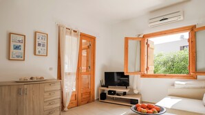 Une pièce moderne et bien éclairée, avec un meuble en bois, un téléviseur posé sur un support, une fenêtre donnant sur un paysage verdoyant et un saladier de fruits sur une table.