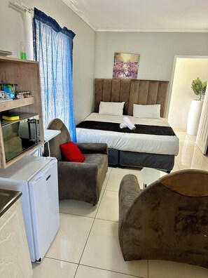 Een hotelkamer met een bed, een klein keukentje, een zithoek met een slaapbank en fauteuil, en een groot raam met gordijnen.