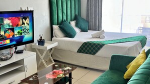 A modern hotel room with a bed, green and white bedding, a green sofa, a small table with a vase, and a TV on a white stand. - Mowetu Guest House (Cape Town)