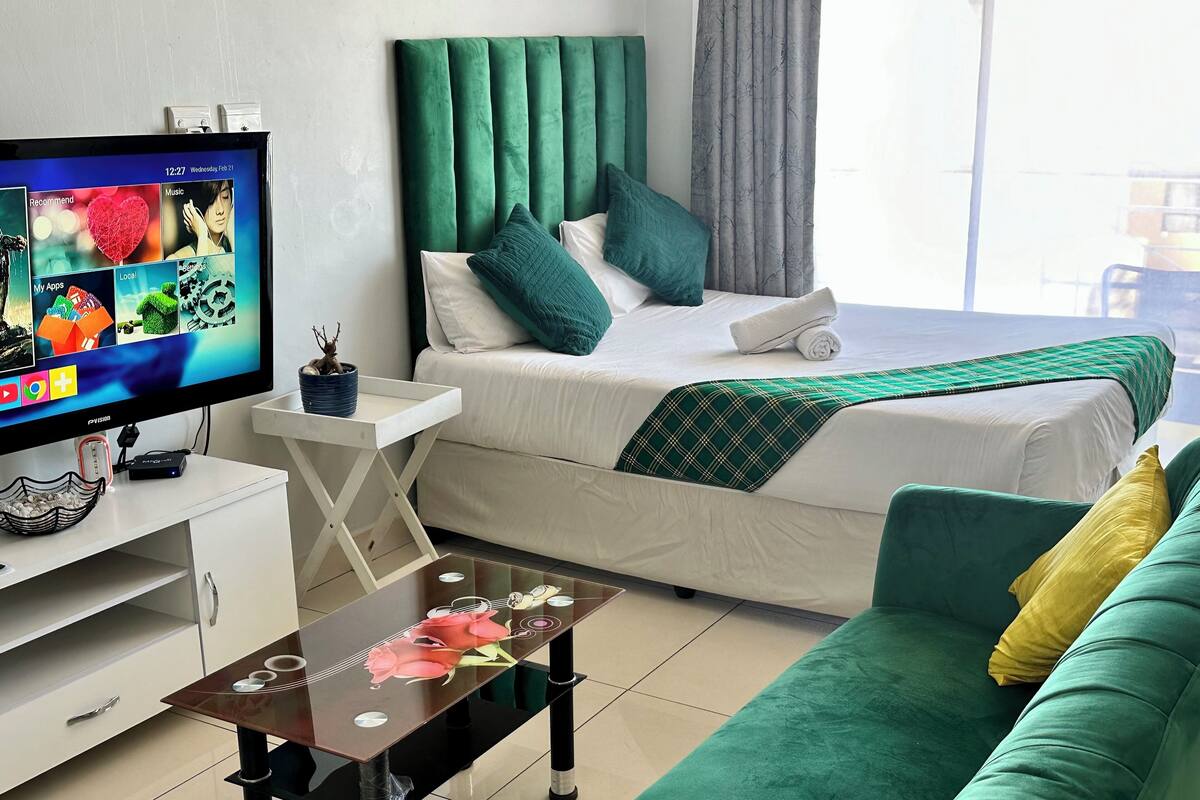 Ein modernes Hotelzimmer mit einem Bett, Bettwäsche in Grün und Weiß, einer grünen Couch, einem kleinen Tisch mit einer Vase und einem Fernseher auf einem weißen Schrank.