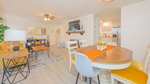 A modern living room with a sofa, coffee table, and dining table. There is a kitchen area with a refrigerator and a TV mounted on the wall. - The Beehive New 4 Bedroom Listing Next to CNU (Newport News)