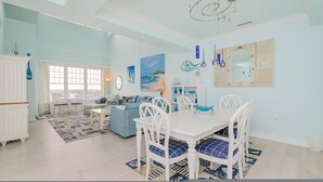 A spacious dining area with a white table and chairs, a blue sofa, and a view of the city through large windows. - 4BR Waterfront Penthouse Pool Views (Virginia Beach)