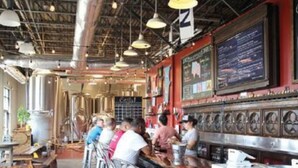 A restaurant interior with high ceilings, exposed beams, and hanging pendant lights. There are metal bar stools and a counter with a menu board. - Ice Ice Baby Downtown Newport News (Newport News)