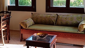A room with a sofa, a wooden coffee table, and a chandelier. There are two windows with a view of greenery outside. - Evinos Village Resort (Nafpaktos)