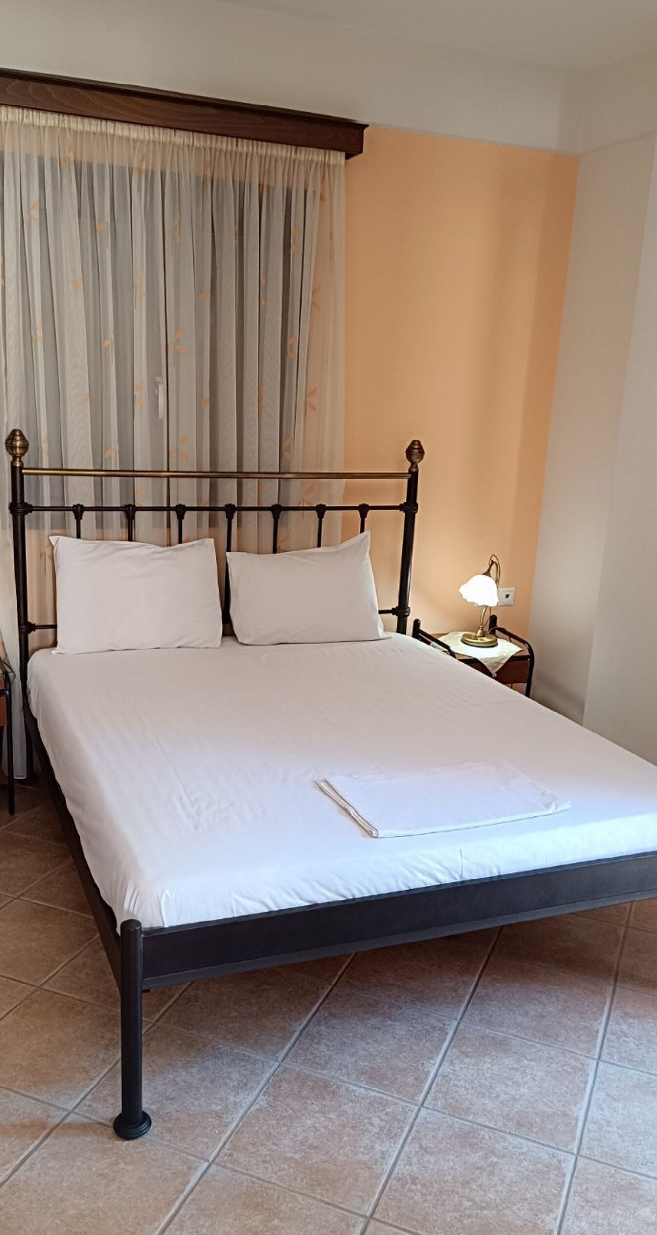 Una cama bien hecha con sábanas y almohadas blancas, una lámpara de noche y una cortina entreabierta.