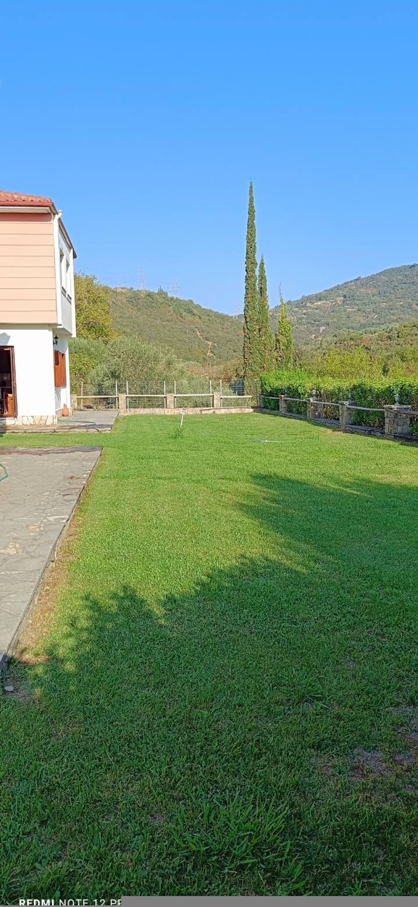 A grassy yard with a building on the left, a fence, and a hill in the background.