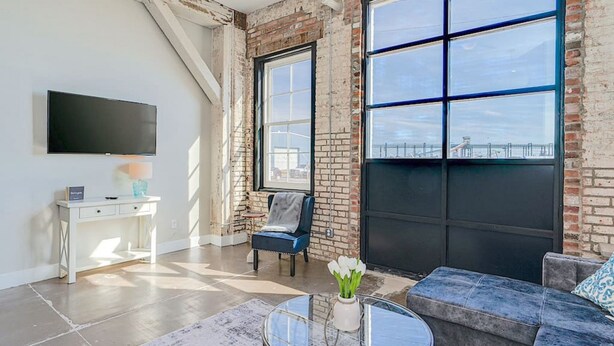 A modern living room with a large window, a flat-screen TV, a white desk, and a blue sofa. - The Speakeasy Downtown Newport News (Newport News)