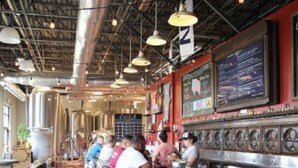 A restaurant interior with high ceilings, exposed ductwork, and a bar area with stools and a counter. - The Safe House Downtown Newport News (Newport News)