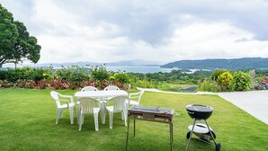 A grassy outdoor area with a barbecue grill, white plastic chairs, and a table. - Ocean View House Nakijin (Nakijin)