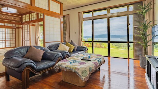 A traditional Japanese-style living room with tatami flooring, a leather sofa, and a large sliding glass door leading to a garden. - Ocean View House Nakijin (Nakijin)
