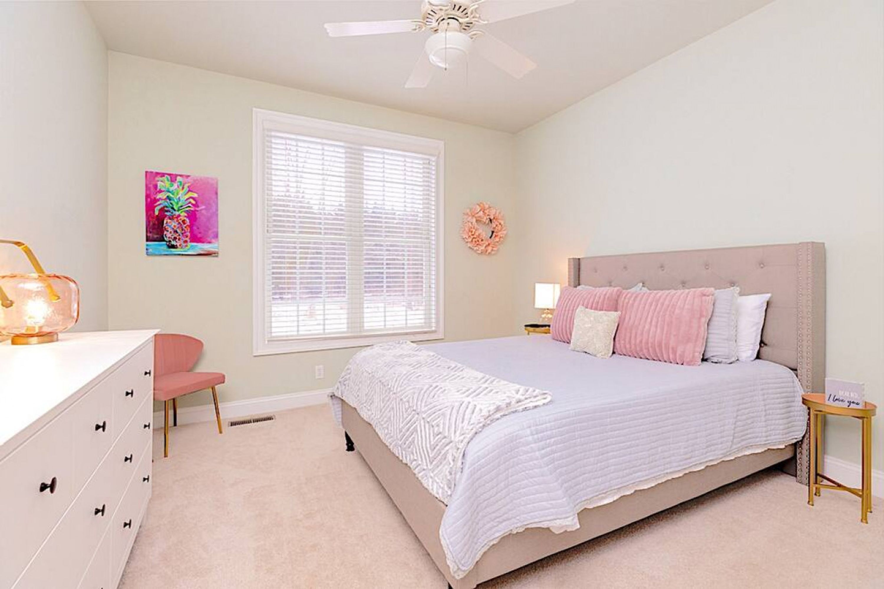A bedroom with a bed, a dresser, a chair, a window with blinds, a ceiling fan, and a wall decoration.